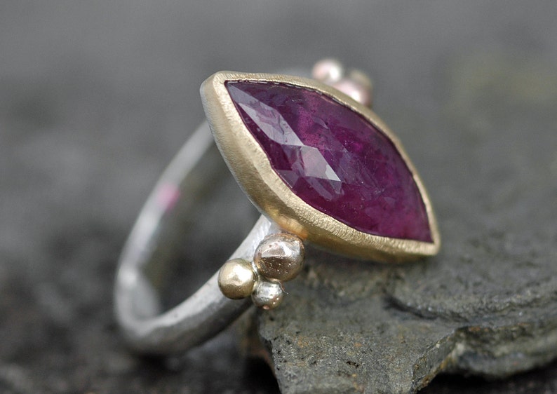 May include: A silver ring with a marquise-shaped ruby gemstone set in a gold bezel. The ring is accented with small gold beads.