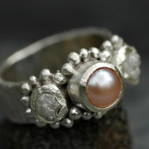 May include: A silver ring with three stones: a pearl, a clear diamond, and a cloudy diamond. The ring is set with a textured band and small silver beads.