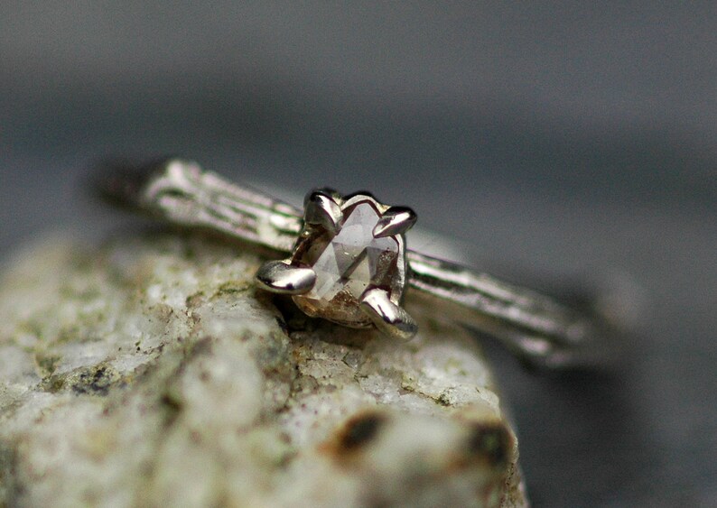May include: A silver ring featuring a delicate twig design and a rough, light brown diamond.