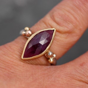 May include: A ring with a marquise-shaped ruby gemstone set in a gold bezel. The band is made of silver and has a beaded design.