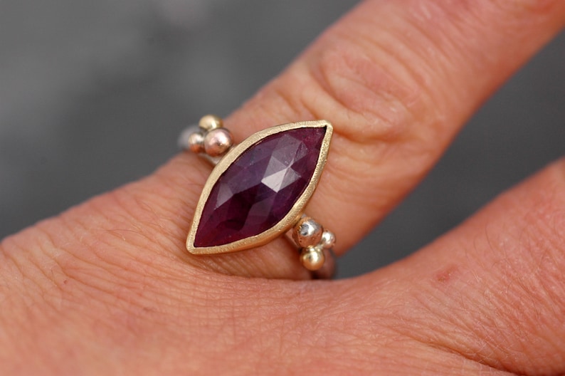 May include: A ring with a marquise-shaped ruby gemstone set in a gold bezel. The band is made of silver and has a beaded design.