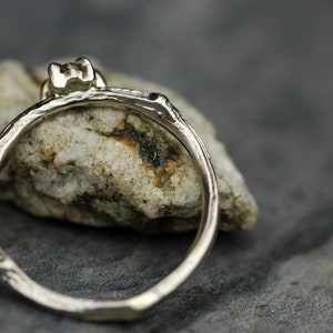 May include: A silver ring with a small, clear gemstone set in a branch-like band. The ring is resting on a piece of rough, white rock.