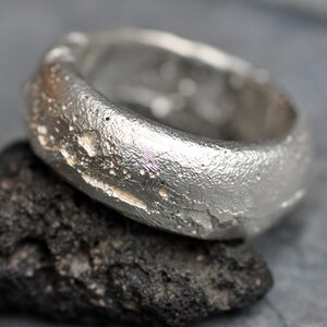 May include: A silver ring with a textured, rough surface. The ring is designed to resemble a piece of rock or a meteorite.