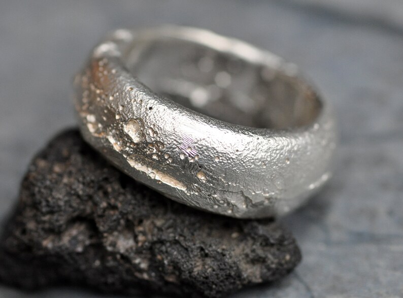 May include: A silver ring with a textured, rough surface. The ring is designed to resemble a piece of rock or a meteorite.