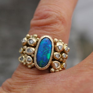 K&ouml;nnte beinhalten: Ein goldener Ring mit einem gro&szlig;en ovalen blauen Opal-Edelstein, umgeben von kleineren Diamanten. Der Ring wird an einem Finger getragen.