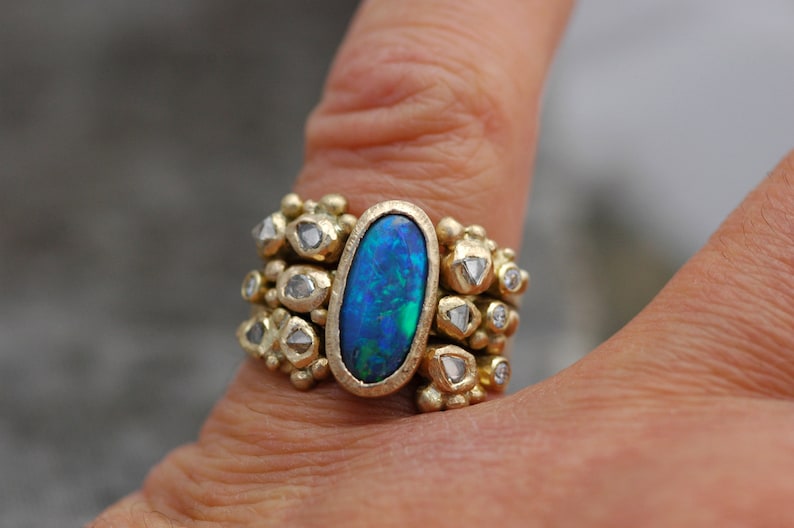 K&ouml;nnte beinhalten: Ein goldener Ring mit einem gro&szlig;en ovalen blauen Opal-Edelstein, umgeben von kleineren Diamanten. Der Ring wird an einem Finger getragen.
