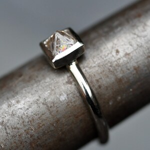May include: A silver ring with a clear, faceted gemstone set in a square bezel. The ring is on a textured metal surface.