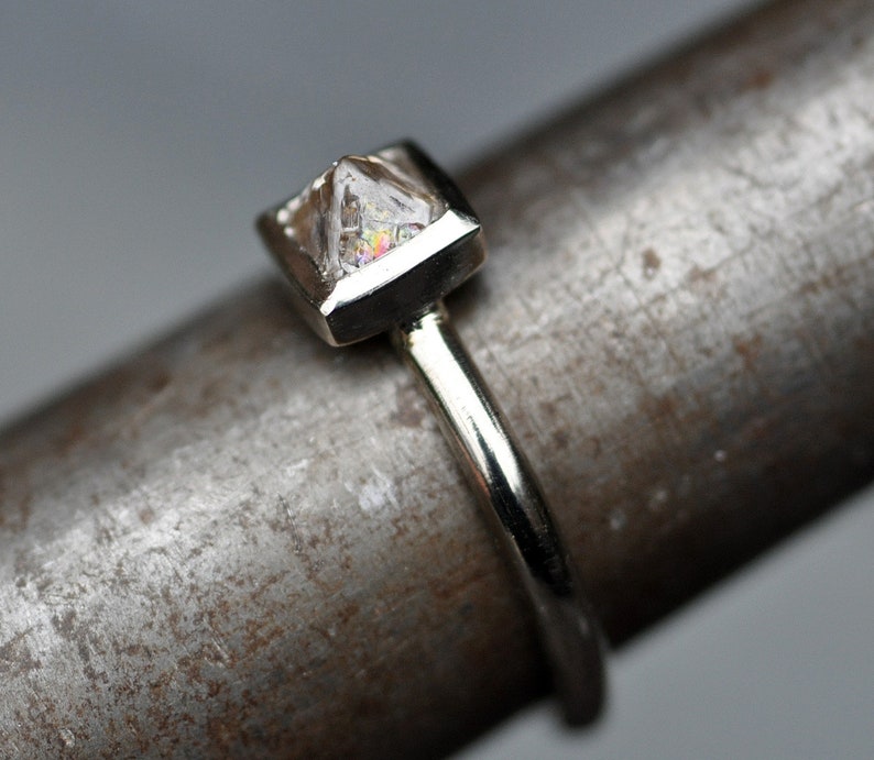 May include: A silver ring with a clear, faceted gemstone set in a square bezel. The ring is on a textured metal surface.