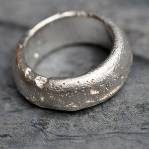 May include: A silver ring with a textured, rough surface. The ring is wide and has a rounded shape.
