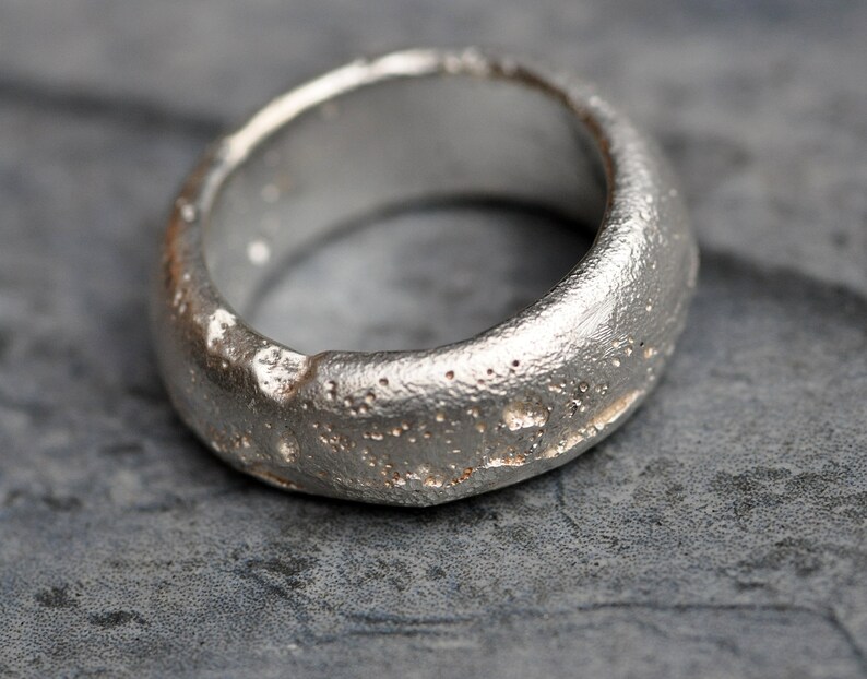 May include: A silver ring with a textured, rough surface. The ring is wide and has a rounded shape.