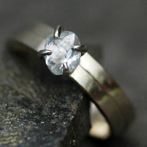 May include: A silver ring with a clear, faceted gemstone set in a prong setting. The ring is on a dark gray background.