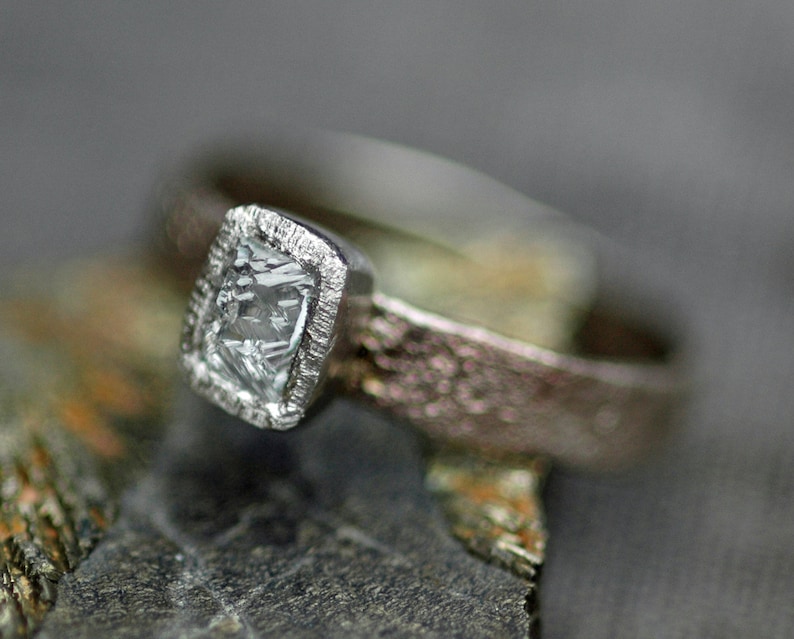 May include: A silver ring with a textured band and a square-shaped clear gemstone set in a silver bezel.