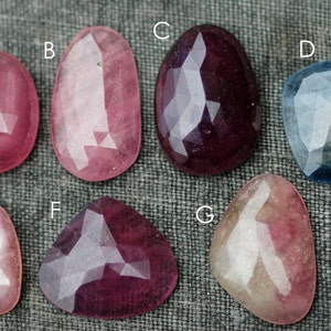 May include: Seven faceted gemstones in various colors, including pink, red, blue, and white. The gemstones are arranged in a row on a gray fabric background.