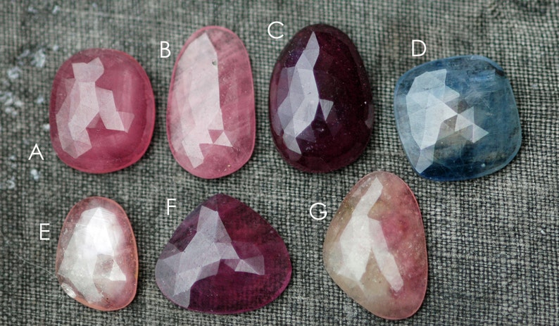 May include: Seven faceted gemstones in various colors, including pink, red, blue, and white. The gemstones are arranged in a row on a gray fabric background.