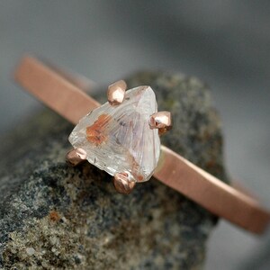 May include: A delicate rose gold ring featuring a raw, clear crystal gemstone. The crystal has internal orange and brown inclusions and is held in place by four rose gold prongs. The band is a simple, thin design.