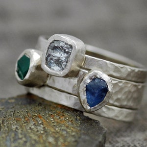 May include: Three silver rings with different colored gemstones. The top ring has a clear, faceted gemstone. The middle ring has a green gemstone. The bottom ring has a blue gemstone.