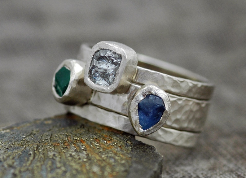 May include: Three silver rings with different colored gemstones. The top ring has a clear, faceted gemstone. The middle ring has a green gemstone. The bottom ring has a blue gemstone.