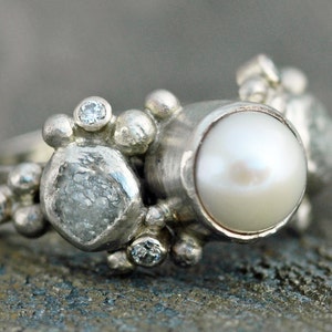 May include: A silver ring with a large white pearl, a rough-cut diamond, and smaller diamonds set in a cluster.