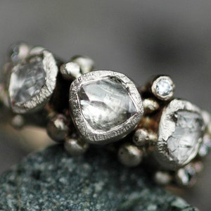 May include: A silver ring with multiple clear, faceted gemstones set in a cluster. The ring is made of metal and has a textured finish.