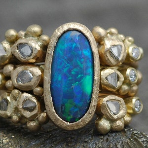 K&ouml;nnte beinhalten: Ein goldener Ring mit einem gro&szlig;en ovalen Opal-Edelstein, umgeben von kleineren Diamanten. Der Ring hat eine strukturierte Band und ein einzigartiges, organisches Design.