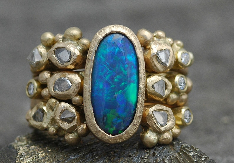 K&ouml;nnte beinhalten: Ein goldener Ring mit einem gro&szlig;en ovalen Opal-Edelstein, umgeben von kleineren Diamanten. Der Ring hat eine strukturierte Band und ein einzigartiges, organisches Design.