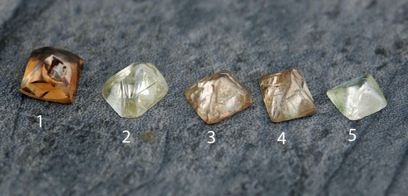 May include: Five different cut diamonds, numbered 1 through 5, are displayed on a gray textured surface. The diamonds are various shades of brown and yellow.