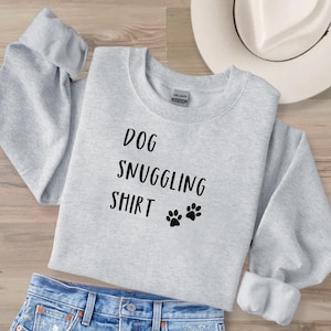 May include: A pale grey sweatshirt featuring the text "DOG SNUGGLING SHIRT" in black, accompanied by two paw prints. The sweatshirt is displayed with blue denim jeans and a white hat.