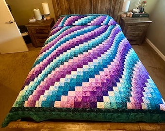 May include: A handmade quilt with a vibrant, geometric pattern in shades of purple, blue, and teal. The quilt is spread across a wooden bed frame, showcasing its intricate design and craftsmanship. The quilt's border is a dark green.