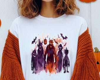 May include: White crew neck t-shirt featuring a watercolor illustration of Halloween monsters in shades of purple, orange, and black. The design includes bats and a jack-o'-lantern. The shirt is part of a Halloween-themed apparel collection.