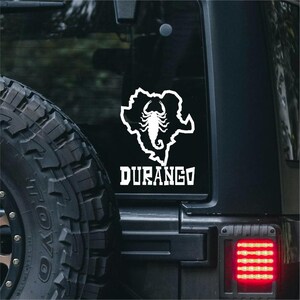 May include: White vinyl decal featuring a scorpion inside a state outline, with the word "DURANGO" below. The decal is affixed to a black vehicle window, near a tire and a red taillight.