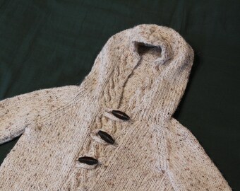 May include: A cream-colored knitted cardigan with a hood and brown buttons. The cardigan has a speckled pattern and is made of a soft, cozy material.