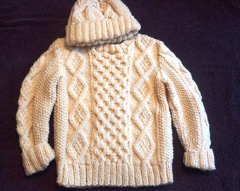 May include: A cream-colored knitted sweater with a matching hat. The sweater has a cable knit pattern and a high neck.