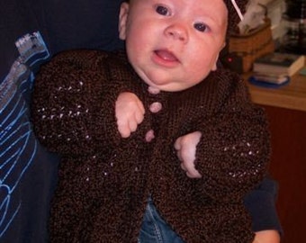 May include: A brown knitted cardigan with a white bow on a brown knitted hat. The cardigan has a button closure and is worn by a child.