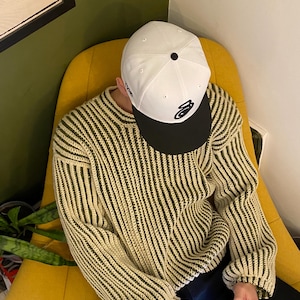 May include: A person wearing a white and black baseball cap with a logo on the front, a green and white striped jumper, and black trousers. The person is sitting in a yellow chair and holding a smartphone.