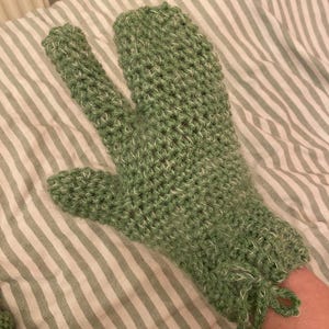 Op de afbeelding: Een groene gehaakte want met een apart gedeelte voor de duim. De want is gemaakt van getextureerd garen met witte vlekjes. De manchet heeft een trekkoordsluiting. De want ligt op een gestreepte stof.