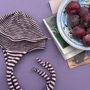 Puede incluir: Un gorro de punto con un patrón de rayas marrones y rosa claro, con un ala ancha y dos lazos trenzados largos que cuelgan. El gorro está colocado junto a un cuenco de cerámica lleno de ciruelas rojo oscuro y una revista con texto visible.