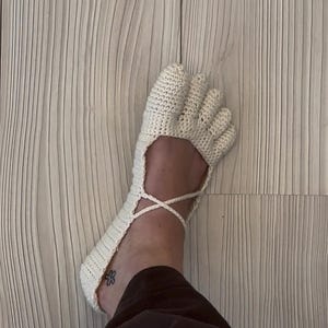 Può includere: Un calzino a dita lavorato all'uncinetto a mano in color bianco sporco, dotato di scomparti individuali per ogni dito e un delicato dettaglio con lacci incrociati che avvolge la parte superiore del piede. Il tessuto ha un aspetto intrecciato.