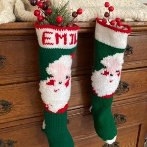 May include: Two green and red knitted Christmas stockings with Santa Claus faces and white fur trim. One stocking has the name "EMIL" knitted in red on the cuff.