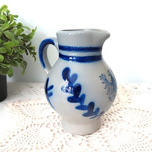 May include: A small, gray ceramic pitcher with a blue and white design. The pitcher has a handle and a spout. It is decorated with a blue and white floral pattern and a blue and white eagle.