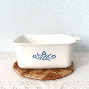 May include: A rectangular, off-white casserole dish with a blue floral design. The dish has two small handles and is resting on a round, woven trivet. The floral design is centered on the front of the dish.