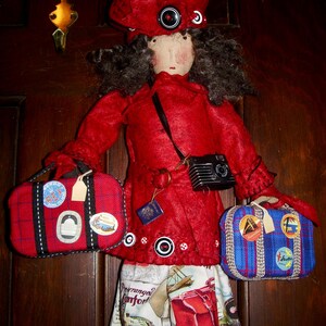 May include: A handmade doll dressed in a red coat, hat, and skirt, holding two travel bags. The doll has a camera around its neck and a passport. The bags are red and blue with travel stickers. The skirt has a travel-themed print.