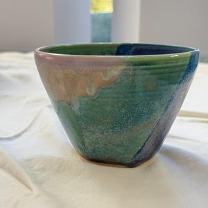 May include: A ceramic bowl featuring a tapered shape and a glossy glaze with blended colors of deep forest green, muted lavender, and earthy tan. The surface displays a subtle speckled texture throughout the exterior and interior walls.