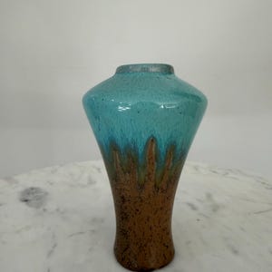 May include: A ceramic vase featuring a wide, tapered body with a glossy turquoise glaze on the upper portion that drips down into a textured, matte brown base. The vessel has a narrow, circular opening at the top and a smooth, rounded silhouette.