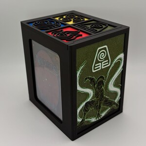 Peut inclure: Une boîte de rangement rectangulaire noire pour cartes avec un panneau latéral illustré d'un personnage en position d'arts martiaux et d'un symbole de terre. Le couvercle présente quatre icônes carrées représentant les éléments eau, feu, air et terre.