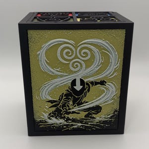 May include: Black square box featuring a yellow-green panel with a white and gray swirling design and a black silhouette of a character in a fighting stance. The top of the box has a decorative design with red, yellow, and blue accents.