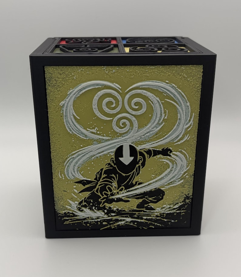 May include: Black square box featuring a yellow-green panel with a white and gray swirling design and a black silhouette of a character in a fighting stance. The top of the box has a decorative design with red, yellow, and blue accents.