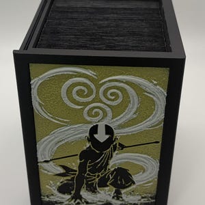 May include: Black card storage box featuring a design of a character in a kneeling pose with a staff, set against a swirling white and yellow background. The box has a black frame and holds a stack of black cards.