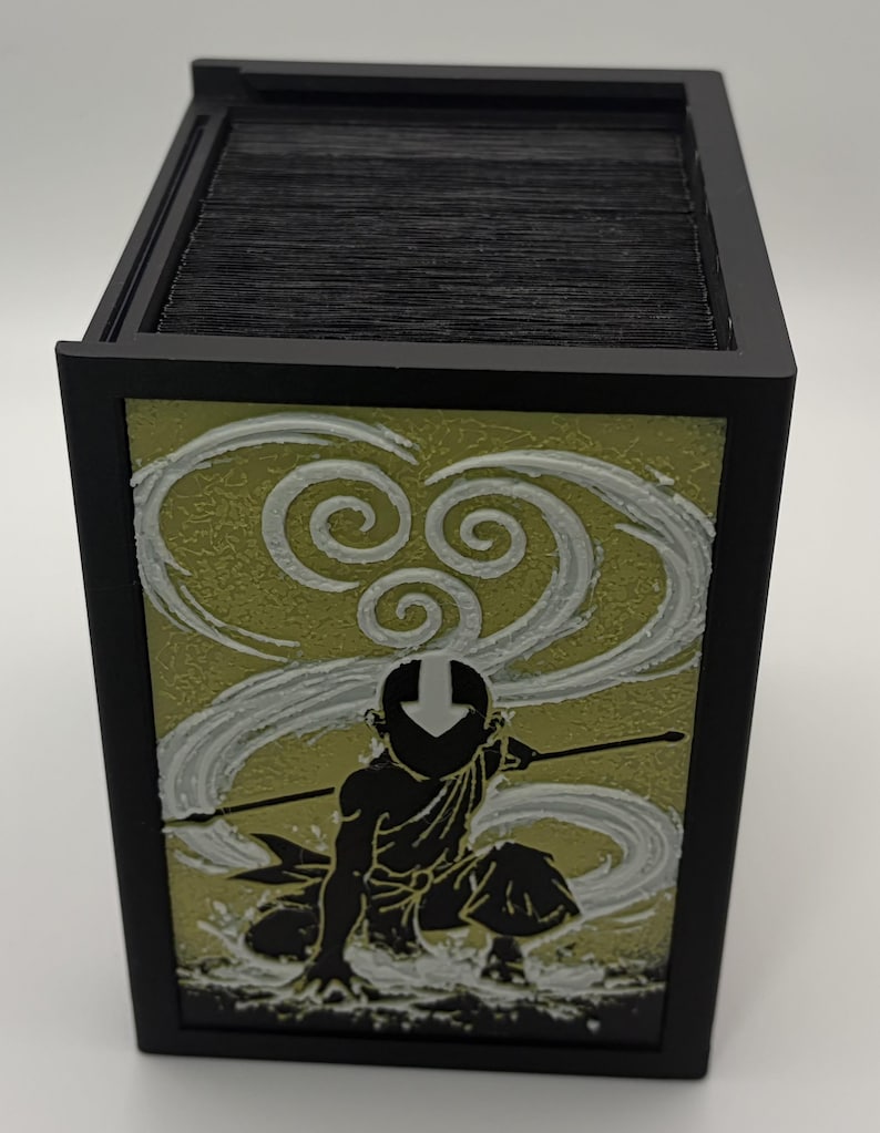 May include: Black card storage box featuring a design of a character in a kneeling pose with a staff, set against a swirling white and yellow background. The box has a black frame and holds a stack of black cards.