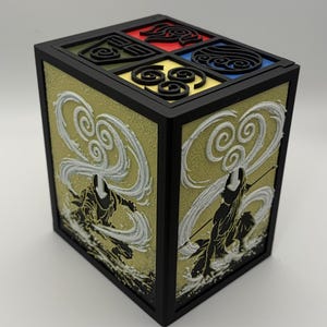May include: A black, square box featuring four elemental symbols in red, blue, yellow, and green. The sides display a textured gold background with white and black figures. The top has a black border.