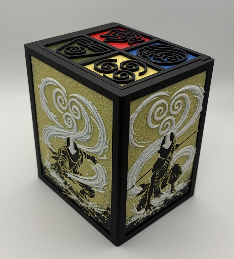 May include: A black, square box featuring four elemental symbols in red, blue, yellow, and green. The sides display a textured gold background with white and black figures. The top has a black border.